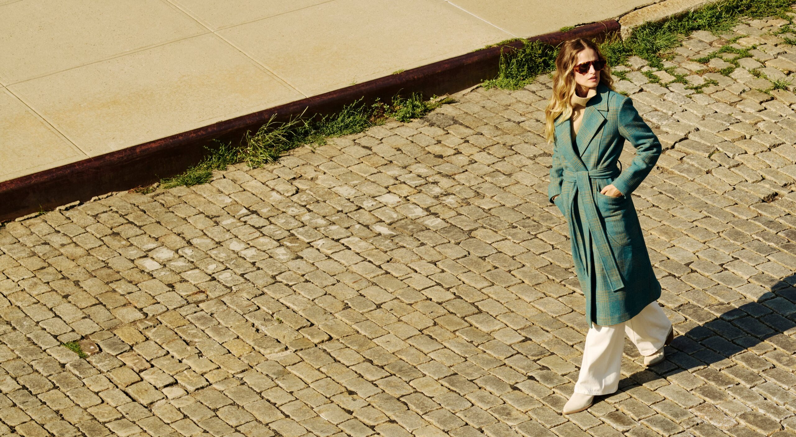 A woman in a teal coat and sunglasses walks on a cobblestone street, with sunlight casting a shadow