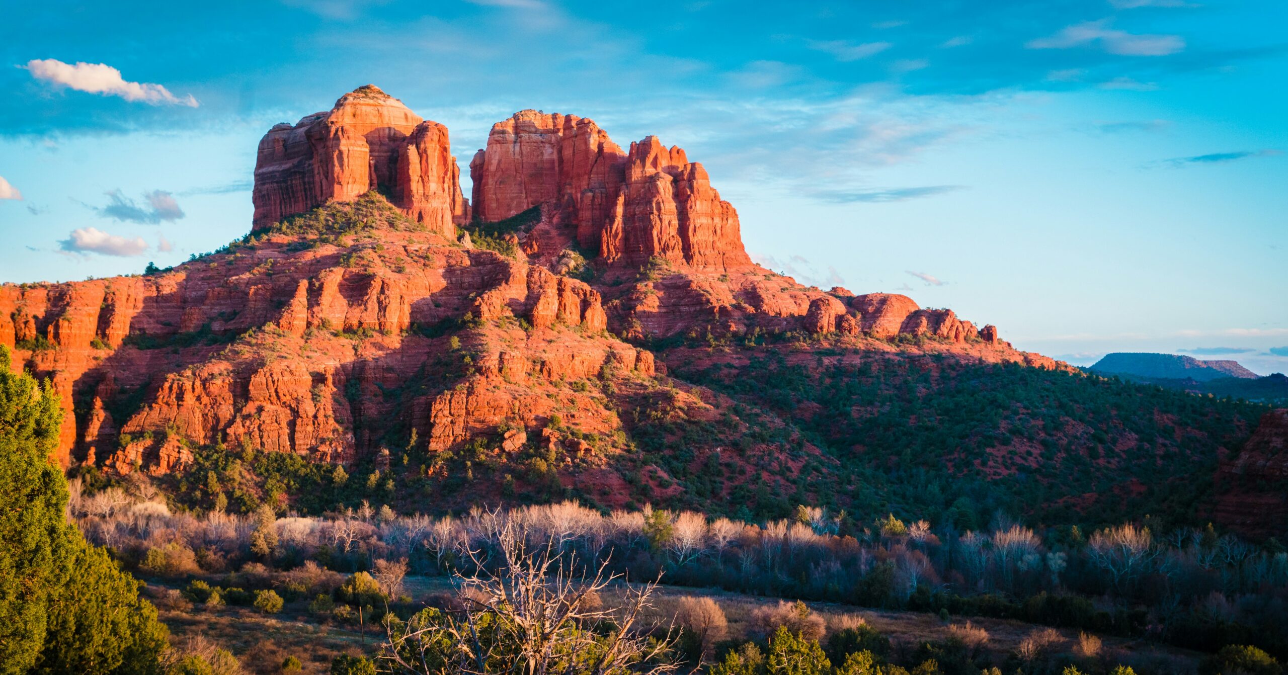 Vibrant red rock formations rise dramatically against a blue sky, with scattered clouds and greenery in the foreground